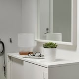 O mică plantă albă în ghiveci FEJKA stă pe un blat de baie, lângă o lampă albă și ziare pliate, afișând un design elegant și minimalist.