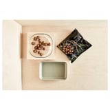 Un vas de cuptor din sticlă transparentă, de formă pătrată, care conține alimente. Este așezat pe o suprafață de lemn cu o tavă dreptunghiulară verde și un produs alimentar ambalat în apropiere.