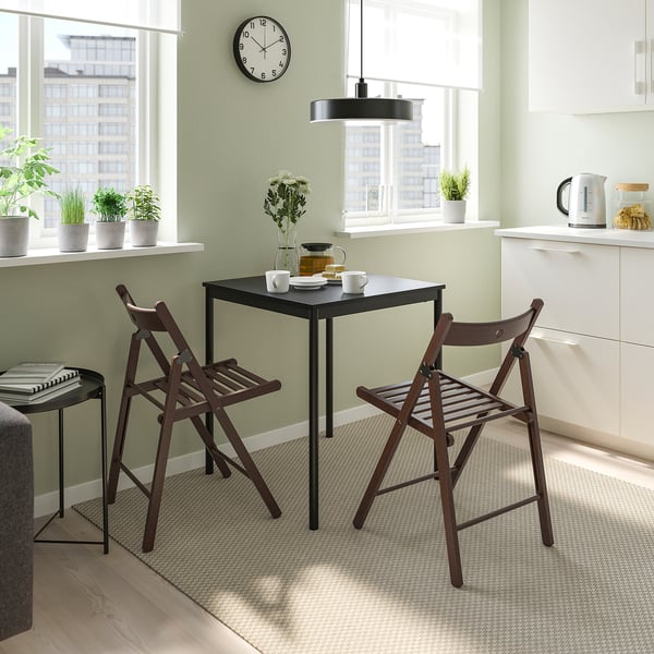 Masă și scaune mici negre FRÖSVI, perfecte pentru bucătării confortabile. Design ușor, pliabil, cu scaune din lemn. Cameră luminoasă cu plante și cafea.