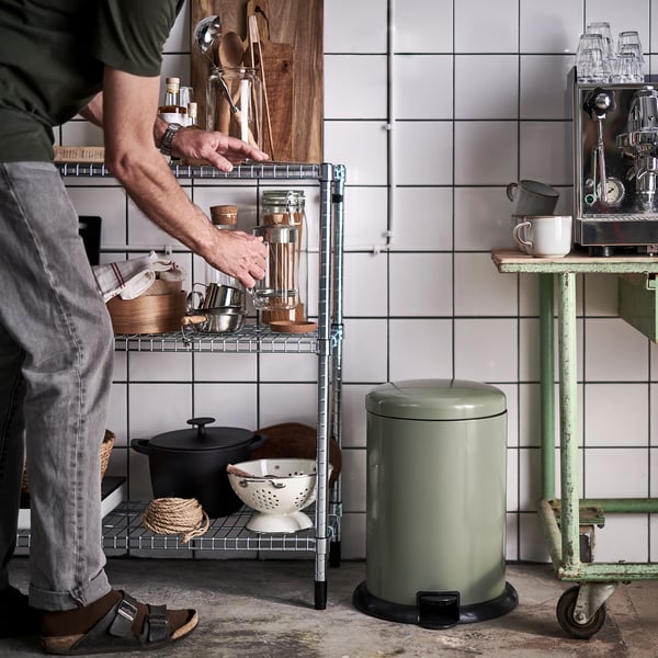 Bărbat care ia obiecte de pe raftul bucătăriei cu recipiente și vase de gătit. Coș verde și aparat de cafea în apropiere.