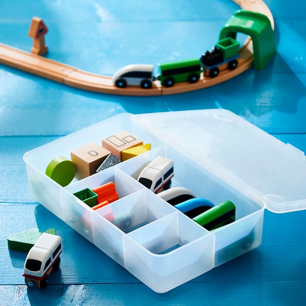 O cutie de depozitare GLIS din plastic transparent cu compartimente conține jucării colorate precum un tren verde, blocuri și vehicule pe o suprafață albastră.