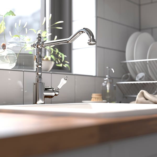 Robinet GLITTRAN argintiu, design elegant, alamă. Gura de scurgere înaltă, lângă chiuvetă cu vase și plantă.