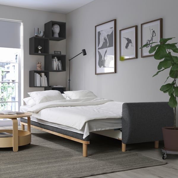 Dormitor modern cu canapea extensibilă gri GRUNNARP, lenjerie de pat albă, artă de perete, stânga: măsuță de cafea cu reviste, dreapta: plantă mare verde.