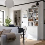 Spațiu de locuit confortabil cu raft de cărți din pin HAVSTA, alb, plin de cărți, decor modern, canapea gri, birou negru, plantă și artă.