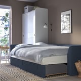 Dormitor modern cu canapea extensibilă gri, lenjerie de pat albă, artă de perete, covor de pluș și masă laterală cu lampă.