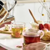 Cești de ceai pe suporturi, ceainic care toarnă ceai, produse de patiserie.