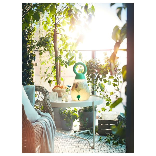 Terasă luminoasă cu o masă rotundă din plasă albă KROKHOLMEN, felinar verde și plante din jur.