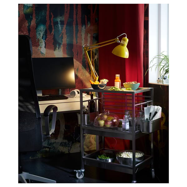 Birou modern de acasă cu cărucior de bucătărie din oțel inoxidabil KUNGSFORS, lampă galbenă, computer, gustări și perdele roșii.