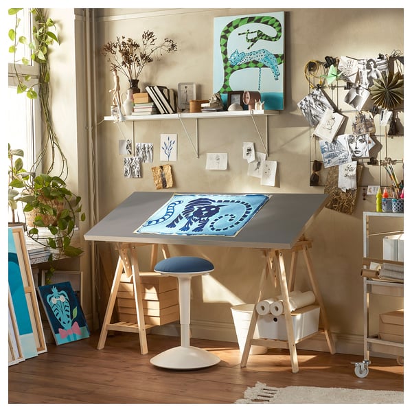 Studio de artă cu birou LAGKAPTEN, lemn deschis, taburet albastru, rafturi cu rechizite de artă, plante, postere și picturi abstracte.