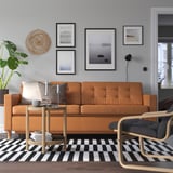 Living modern cu canapea din piele portocaliu-maro, artă, masă laterală, scaun, covor în carouri și plantă.