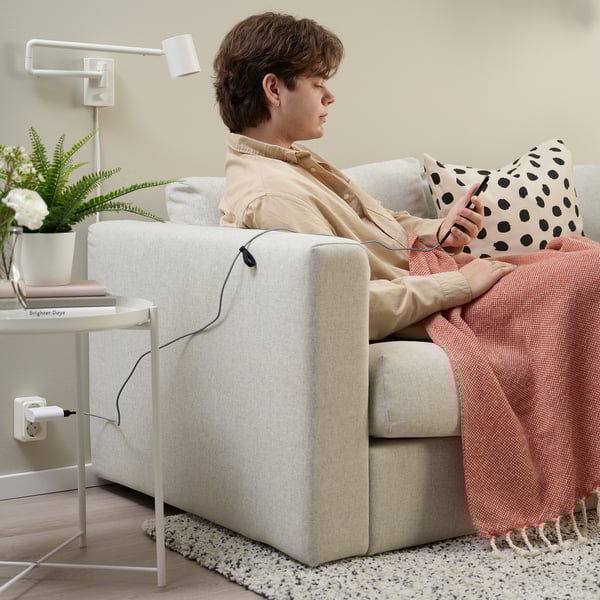 Persoana stă pe canapea bej, folosind un smartphone, înfășurată într-o pătură roșie. O masă laterală albă ține o plantă și o carte intitulată Brighter Days.