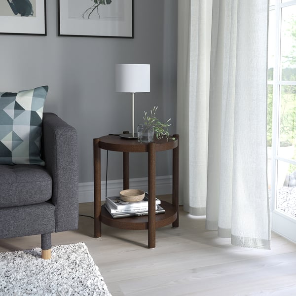 LISTERBY masă laterală în cameră minimalistă cu lampă, plantă, cărți, bol. Lemn închis la culoare, două rafturi, fotoliu gri cu pernă.