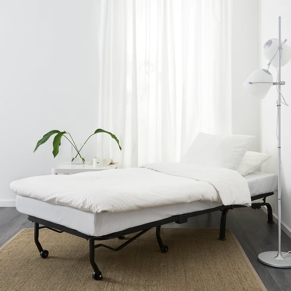Scaun-pat alb modern cu cadru negru elegant. Ideal pentru încăperi compacte. Lampă înaltă, masă laterală cu plantă. Pardoseli deschise, covor bej.