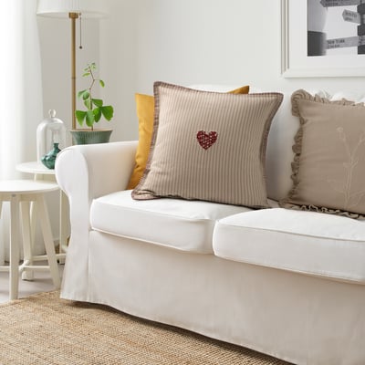 Zona de living cu pernă MÄVINN, inimă roșie pe dungi bej, pe canapea albă cu perne galbene și măsuță laterală cu plante verzi.