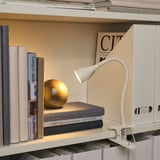 Raft cu cărți și revistă, iluminat de o lampă albă, reglabilă. Un mic decor de glob se sprijină deasupra cărților.