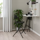 Birou minimalist de acasă: birou alb montat pe perete, scaun pliabil negru. Verdeață, perdele albe. Compact, eficient.