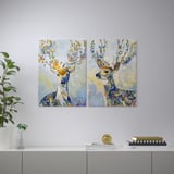 Două tablouri vibrante cu cerbi cu coarne florale atârnă pe un perete deasupra unui raft gri. Raftul conține diverse obiecte decorative.