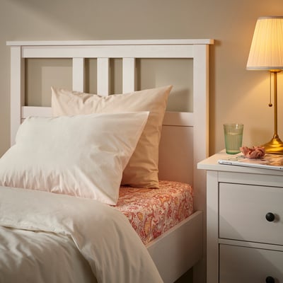 Pat confortabil cu cearșaf paisley RODGERSIA, tăblie albă și perne portocalii. Noptieră cu lampă lângă ea.