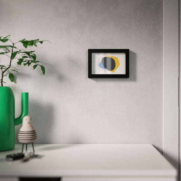 Rama modernă de artă de perete cu design abstract colorat pe un perete gri, afișată deasupra unei mese albe cu o vază verde și o plantă.