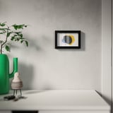 Rama modernă de artă de perete cu design abstract colorat pe un perete gri, afișată deasupra unei mese albe cu o vază verde și o plantă.