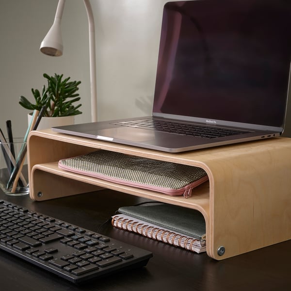 Suport pentru laptop din lemn ROMELSÖN, maro deschis, design curbat, ține laptopul sus, tastatura încape dedesubt, o mică plantă și pixuri în apropiere.