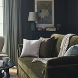 Canapea măslinie cu perne gri, masă laterală neagră cu cană, perdele întunecate, lampă, artă macara.