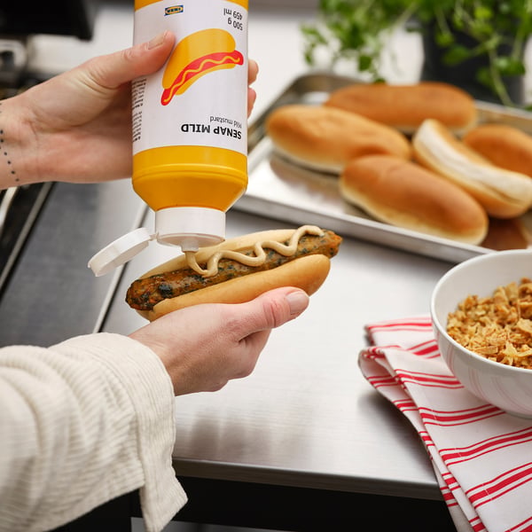 O persoană stoarce muștar galben pe un hot dog. Eticheta sticlei scrie SENAP MILD.