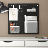 Placă perforată SKÅDIS neagră cu accesorii albe. Include lampă, hârtii, pixuri și coșuri din plasă, montate deasupra biroului alb.