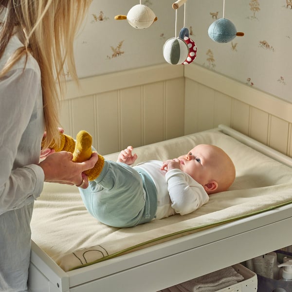 Bebelușul se întinde pe salteaua de schimbat din bumbac biologic. Mama se joacă cu picioarele bebelușului, purtând șosete. Telefonul mobil atârnă deasupra patului.