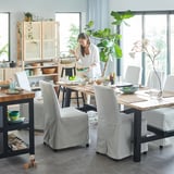 Femeie care servește mâncare în bucătărie modernă cu masă SKOGSTA, scaune gri, rafturi din lemn și plante verzi.