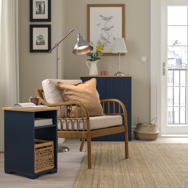 Colț confortabil cu măsuță laterală bleumarin SKRUVBY, scaun de răchită, lampă și artă. Culoarea închisă a meselor contrastează cu decorul deschis, potrivindu-se perfect lângă scaun.