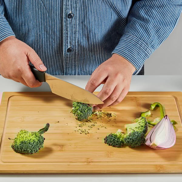 O persoană toacă broccoli pe o tăietură de bambus folosind un cuțit din oțel inoxidabil. Cuțitul taie cu ușurință leguma.