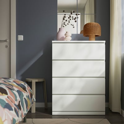 Comodă albă cu 4 sertare în dormitor, design modern cu linii orizontale și mânere integrate. Partea de sus adăpostește lămpile, vazele și lucrările de artă.