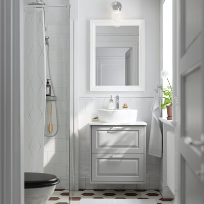 Baie modernă cu chiuvetă albă TÄNNFORSEN, chiuvetă, oglindă montată pe perete, duș din sticlă, gresie cu modele și plante în ghiveci.