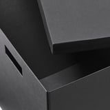 Cutii de carton negre, stivuite cu grijă. Cutia superioară ușor deschisă, arătând o construcție robustă. Material reciclat de înaltă calitate.
