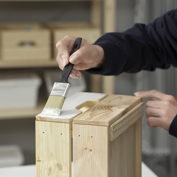 Persoană care perie vopsea albă pe mobilier din lemn într-un atelier. Vopseaua este probabil TRIXIG, care este concepută pentru mobilier interior din lemn.