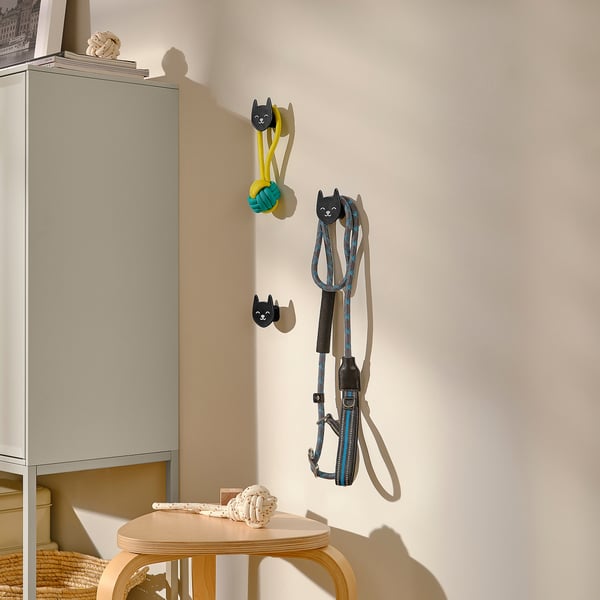 Trei cârlige decorative în formă de pisică atârnă pe un perete, ținând lese și jucării pentru animale de companie, arătând utilizarea practică a depozitului.