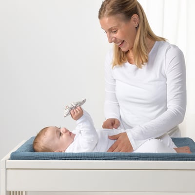 Femeie care schimbă scutecul bebelușului pe covorașul VÄDRA cu husă albastră și bej, folosind jucărie pentru pisici.