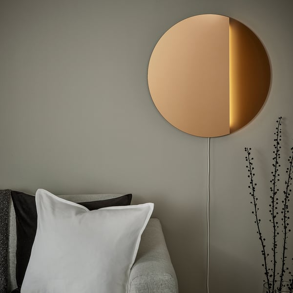 Lampă cu oglindă semicirculară, din bronz, montată sus pe perete, lângă pernele de pe canapea gri deschis. Design modern, minimalist.