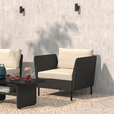 Decor modern de lounge exterior din nuiele neagră, cu perne, măsuță de cafea, pahare de vin și difuzor, așezat pe un perete de beton gri.