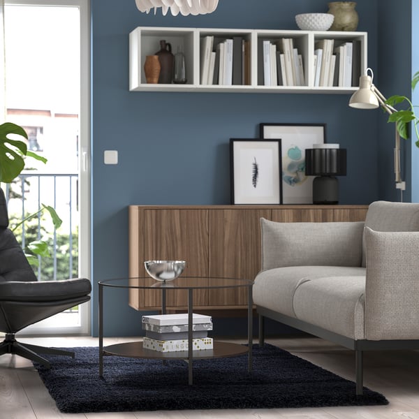 Living confortabil cu covor albastru închis, mobilier modern, masă de sticlă și raft de perete.