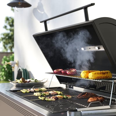 Black outdoor grill with multiple food items cooking. Include adjustable feet, a built-in thermometer, and a warming rack.