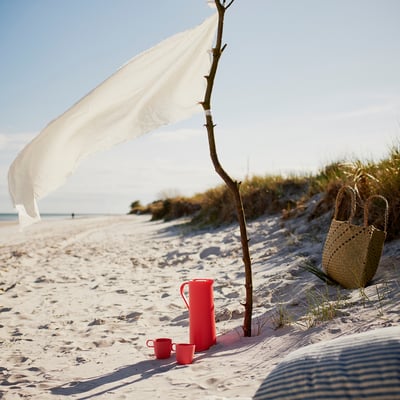 Beach scene with sand, sea, and white cloth draped on stick. Red BEHÖVD thermo flask and two cups on sand, woven basket nearby.