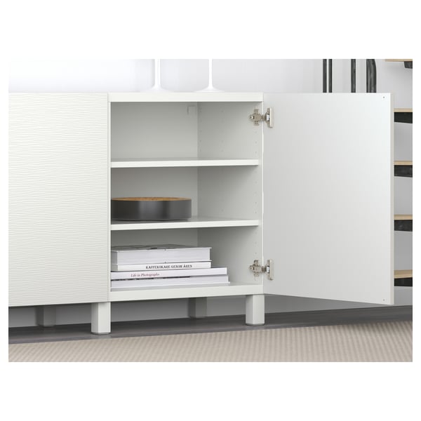 White BESTÅ storage unit with shelves, one door open revealing books and decor. Soft-close hinges visible.