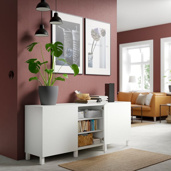 White BESTÅ storage unit with adjustable shelves, displaying books and a plant, set against a red wall with framed art.