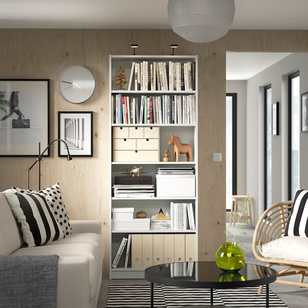 Modern living room with white couch, patterned pillows, wooden bookshelf filled with books, white clock, and a black coffee table with a green vase.