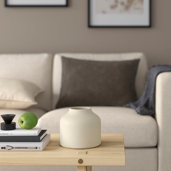 Cosy living room with cream sofa, throw pillow, wooden coffee table, BLODBJÖRK vase, books, apple, decorative item, and blanket.
