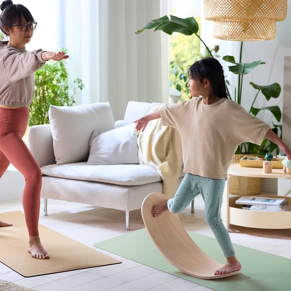 Two people on balance boards, one standing, one in a rocking pose. Indoors, near a white couch.