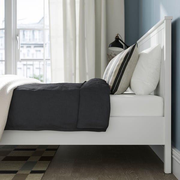 A white bed frame with a grey comforter and striped pillows, featuring slatted base and a tall headboard. Underbed space for storage is visible.