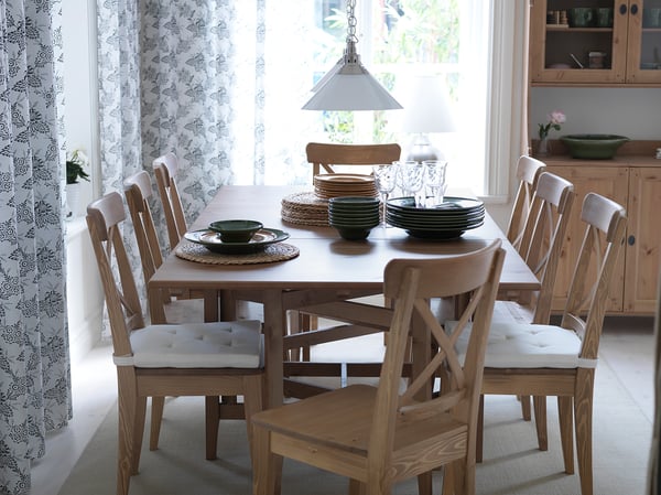 Bright dining room with INGOLF table, six wooden chairs, white cushions, green dishes, floral curtains, and wooden cabinet.